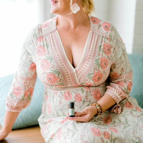 A person sitting on a couch, holding a 10ml essential oil bottle, with a floral patterned dress and a smile on their face.