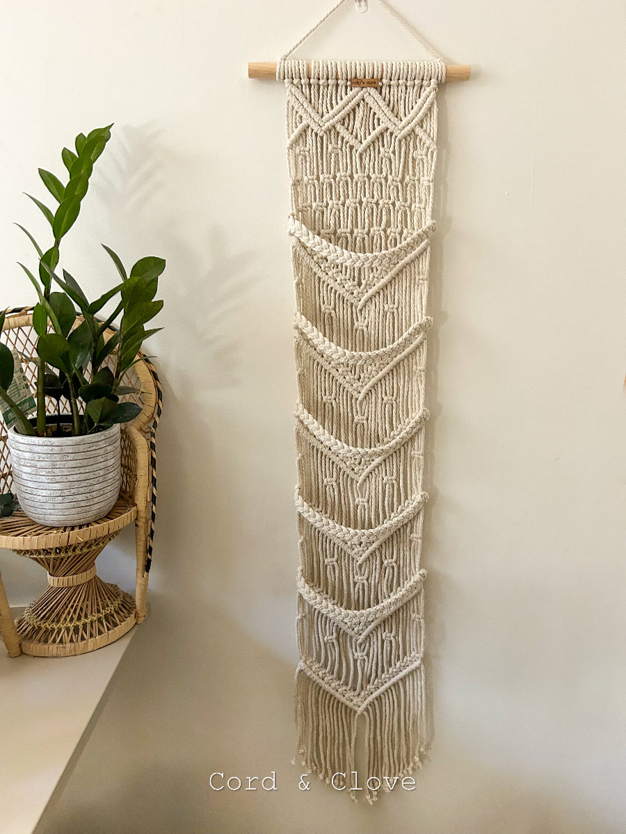 Macrame wall hanging cap storage on a white wall with a plant on a small shelf below.