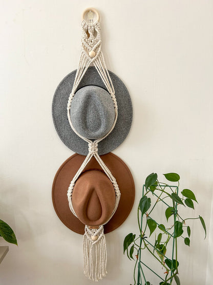 A beaded macrame double hat hanger in off white, hanging on a wall with a plant to the side.