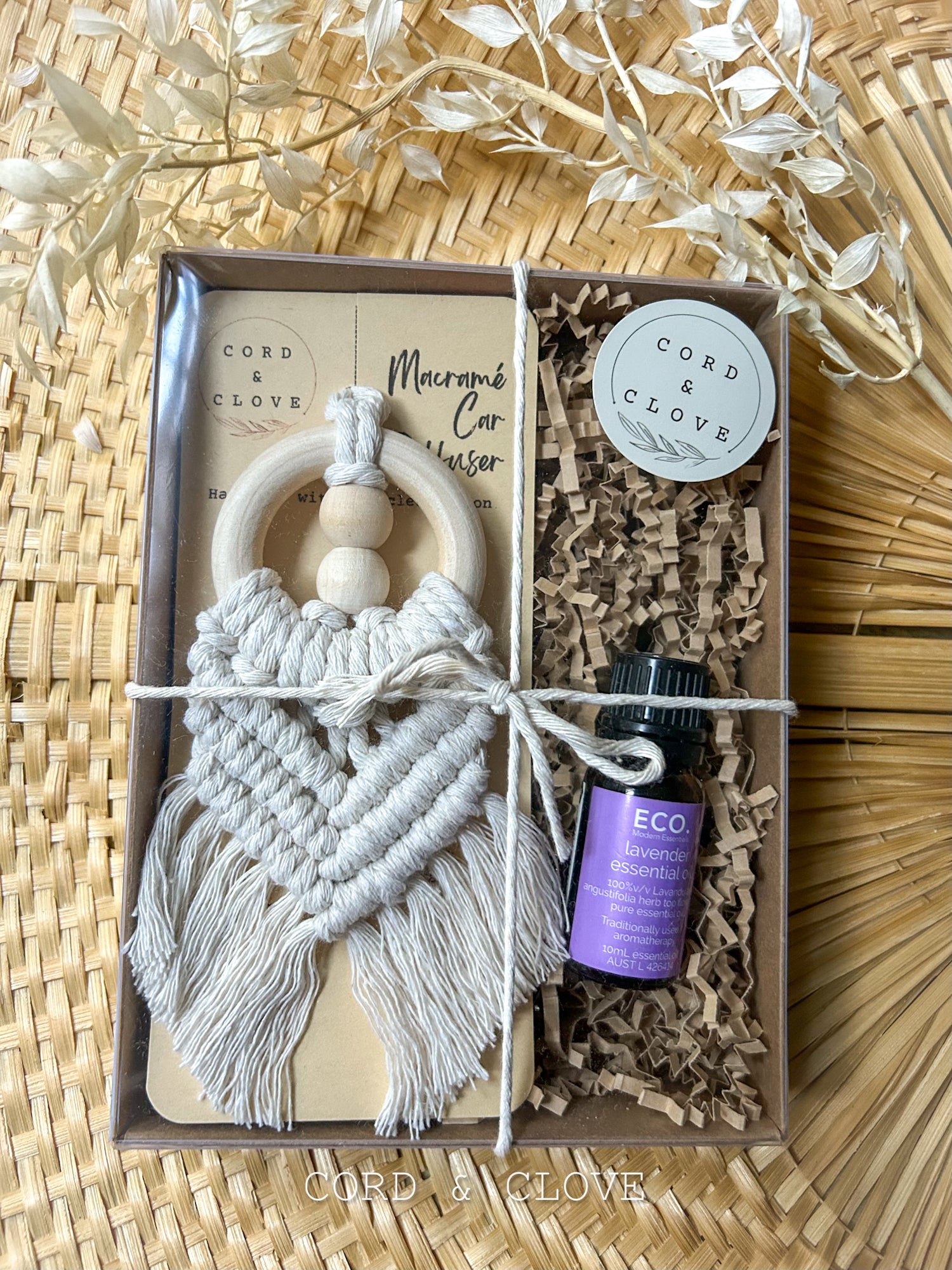 Macrame car hanging kit with essential oil bottle in a cardboard box on a woven surface.