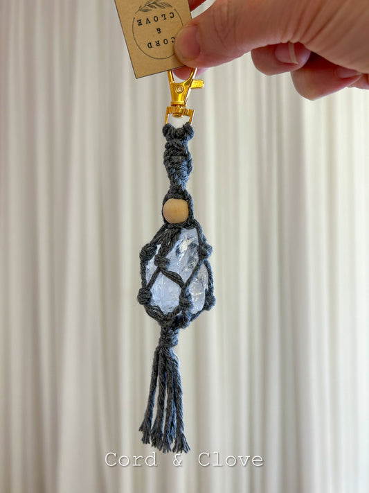Macrame keychain with clear quartz crystal and wooden bead held by a hand against a neutral background.