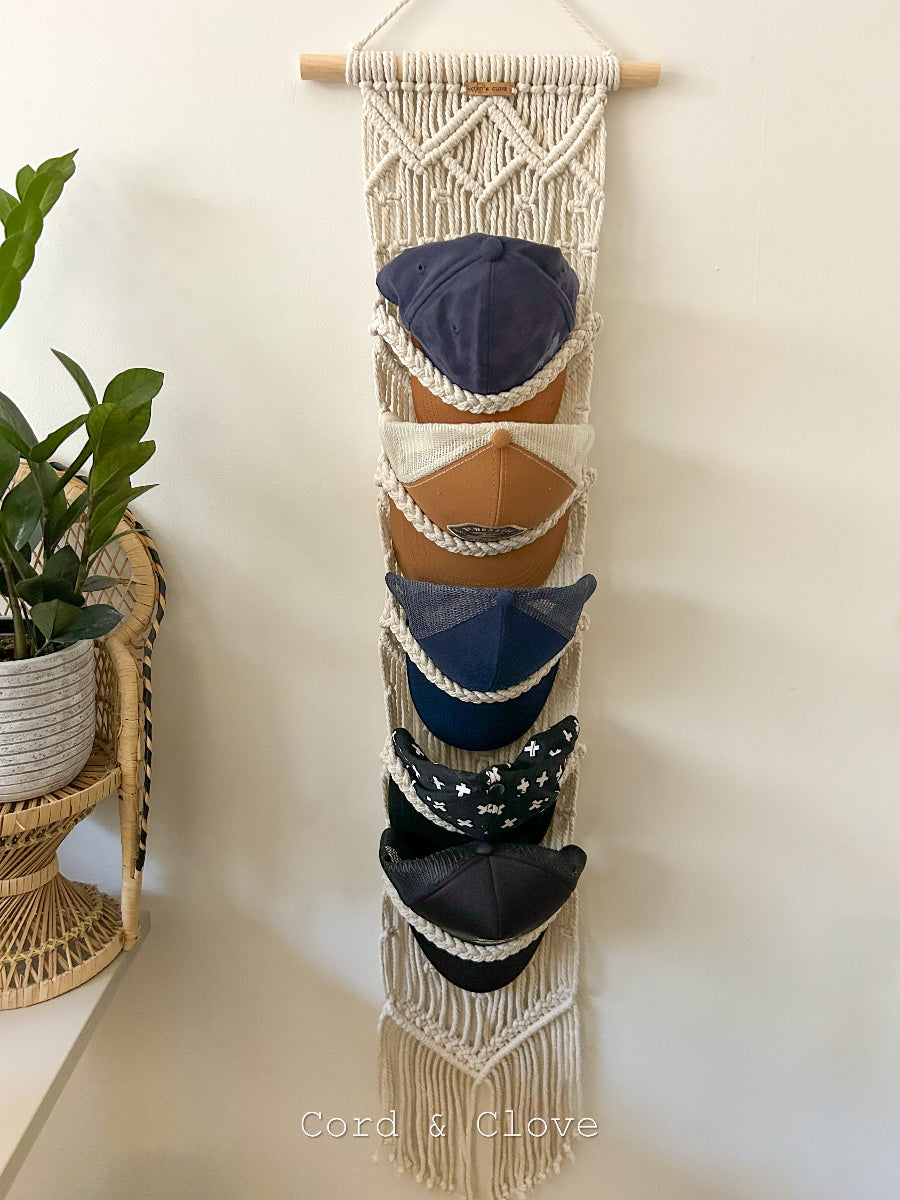 Macrame hat rack with baseball caps hanging on a white wall, featuring the brand 'Cord & Clove'.