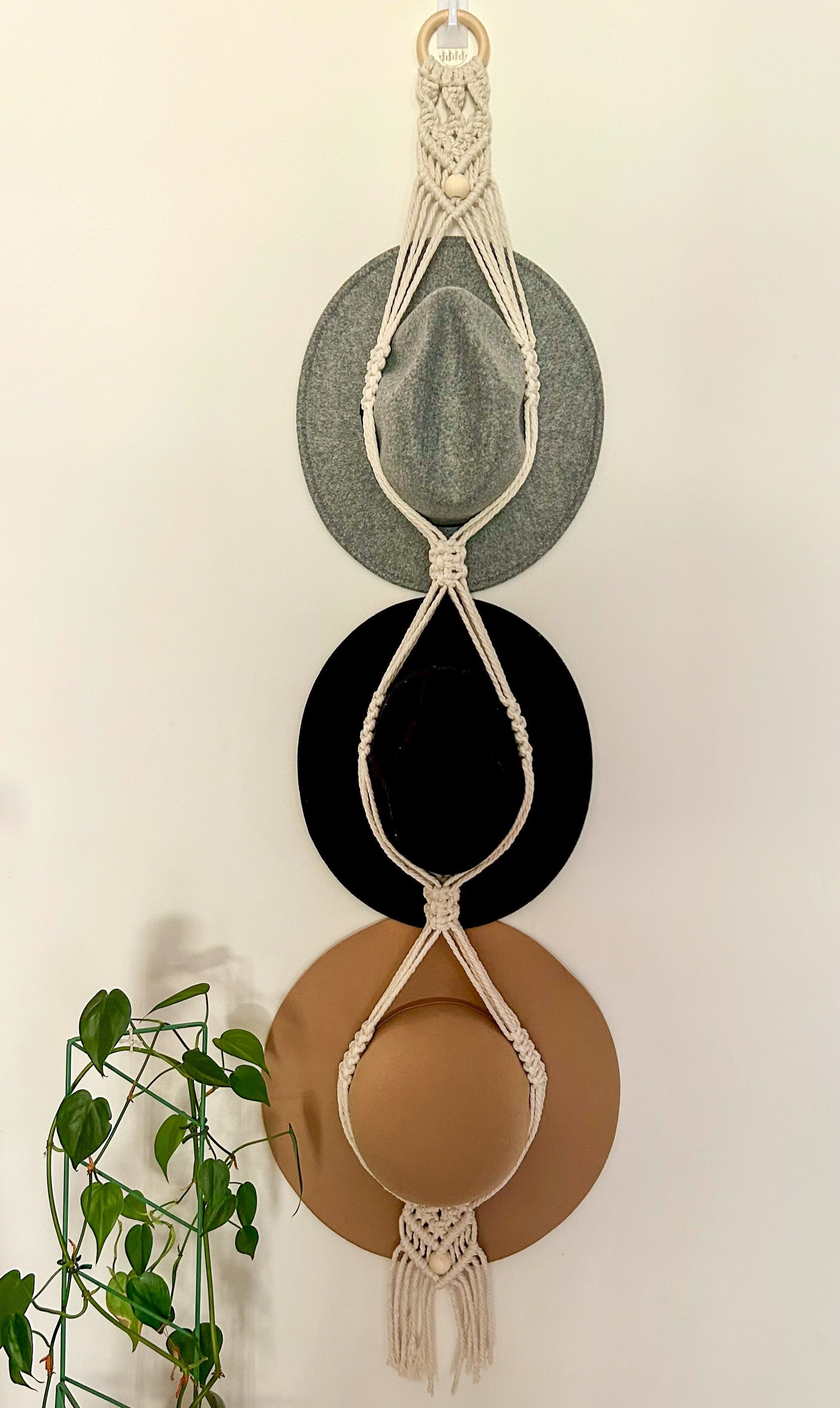 A triple hat hanger with macrame detailing, displaying three brimmed hats in beige, black, and brown colors, hanging against a white wall with a wooden ring and green foliage to the side.