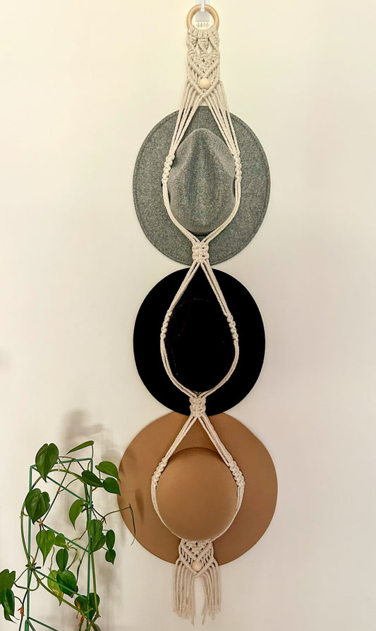 A triple hat hanger with macrame detailing, displaying three brimmed hats in beige, black, and brown colors, hanging against a white wall with a wooden ring and green foliage to the side.