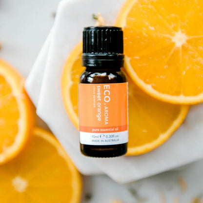 A bottle of Eco Modern Essentials sweet orange essential oil placed on a surface with several sliced oranges and leaves, with a background of uncut oranges.