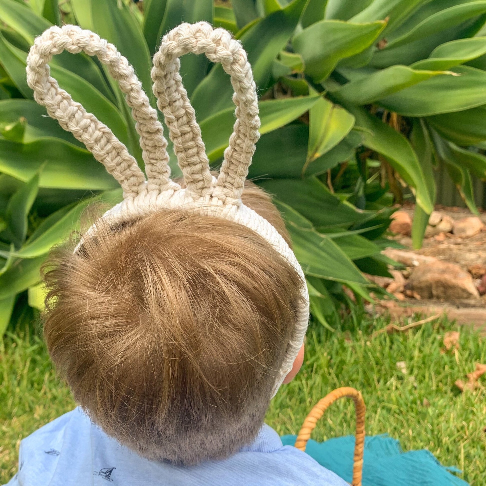 A child wearing an off white macrame headband shaped like a bilby or bunny ears, with a garden in the background.